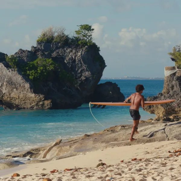 A surfer carrying a longboard across rocks toward the sea.