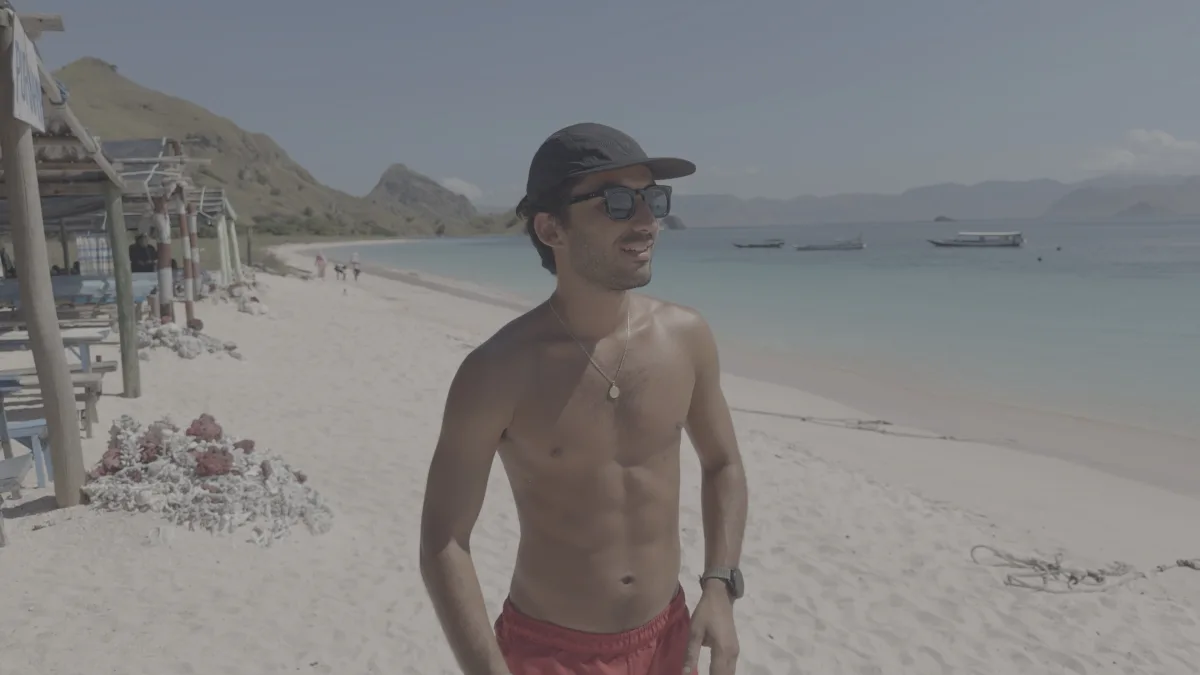 A man on a white-sand beach with turquoise water behind him, in flat Apple Log.